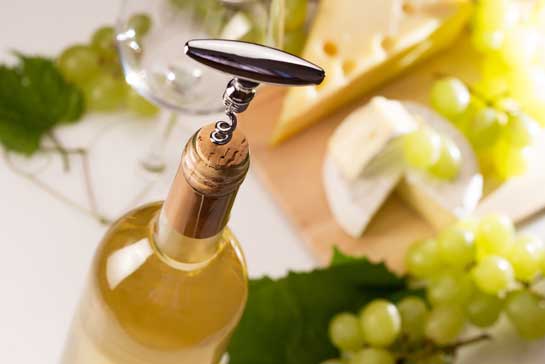Corkscrew in cork of wine bottle; cheese platter and grapes in background 